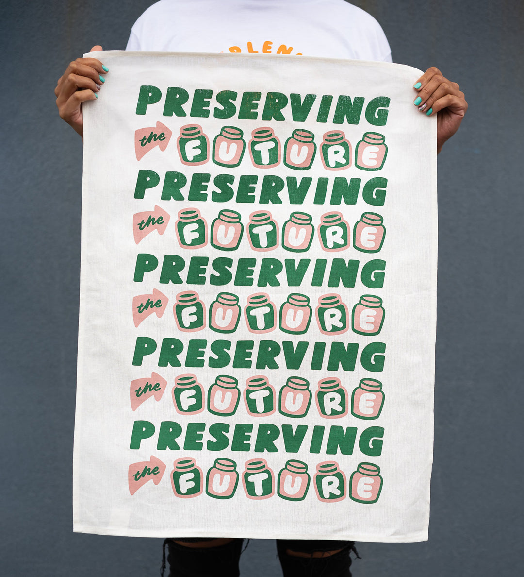 hands holding white linen and cotton blend tea towel with five rows of green and pink text staying preserving the future