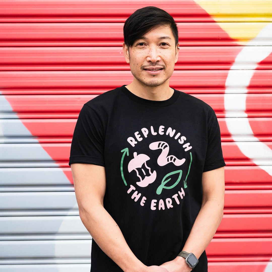 smiling man in front of colourful wall wearing black organic cotton shirt with cute pink and green design saying replenish the earth around an apple, worm and leaf