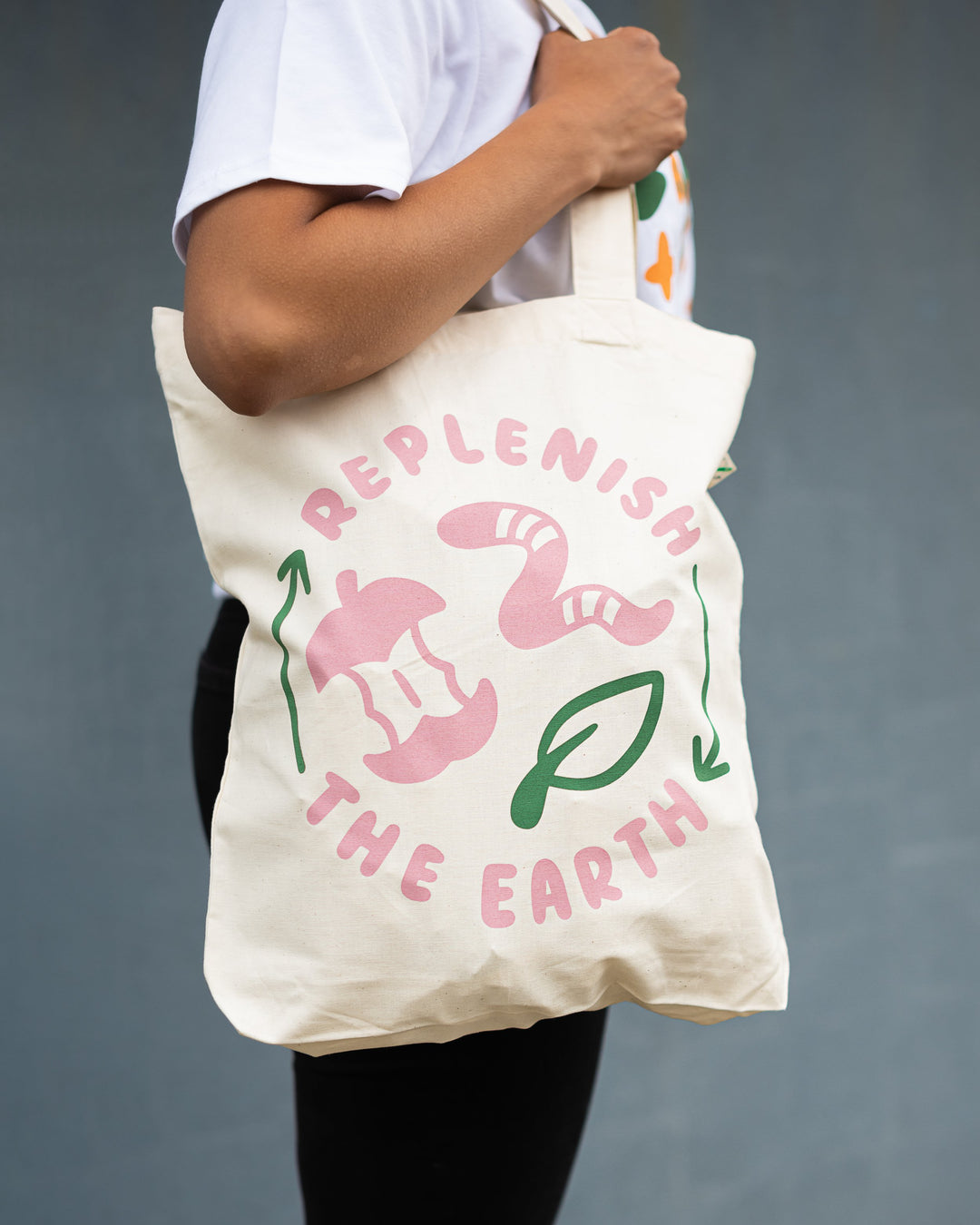 arm holding organic white cotton tote with pink and green design saying replenish the earth with apple, worm and leaf