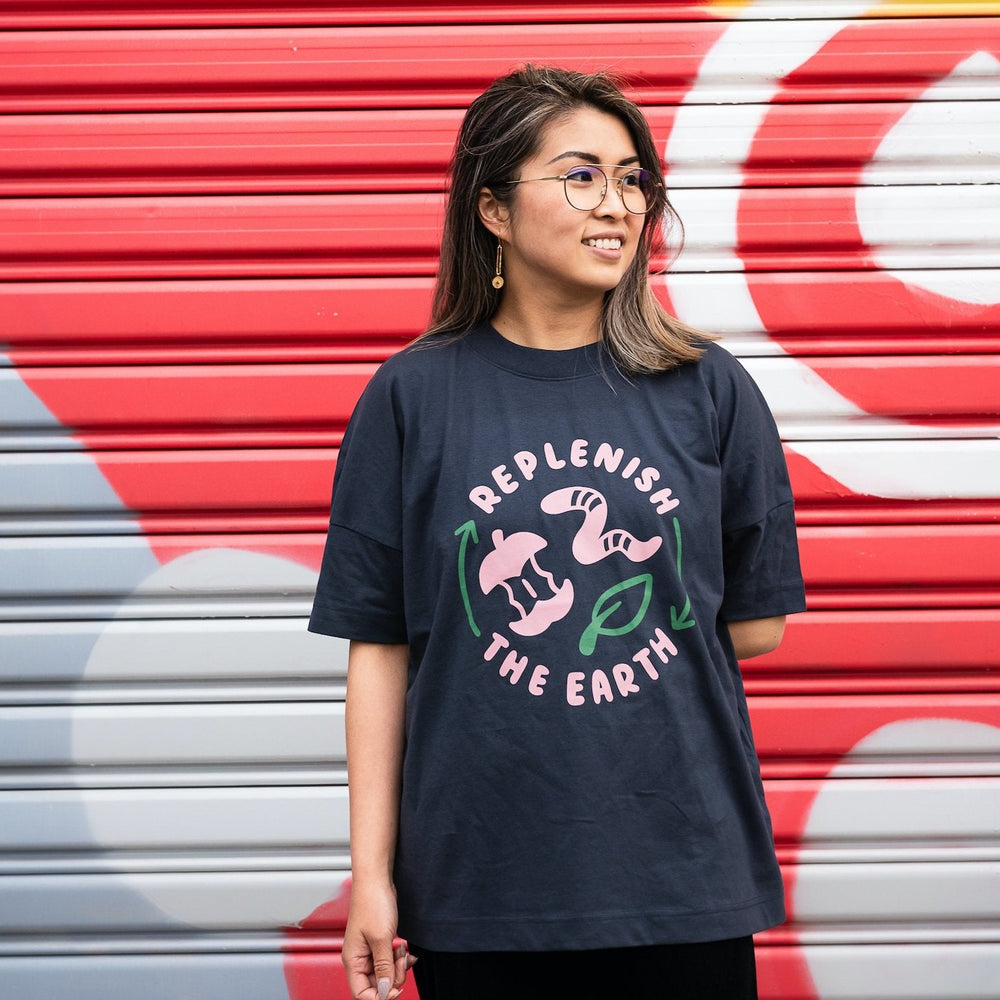 smiling woman in front of colourful wall wearing oversized grey organic cotton shirt with cute pink and green design saying replenish the earth around an apple, worm and leaf