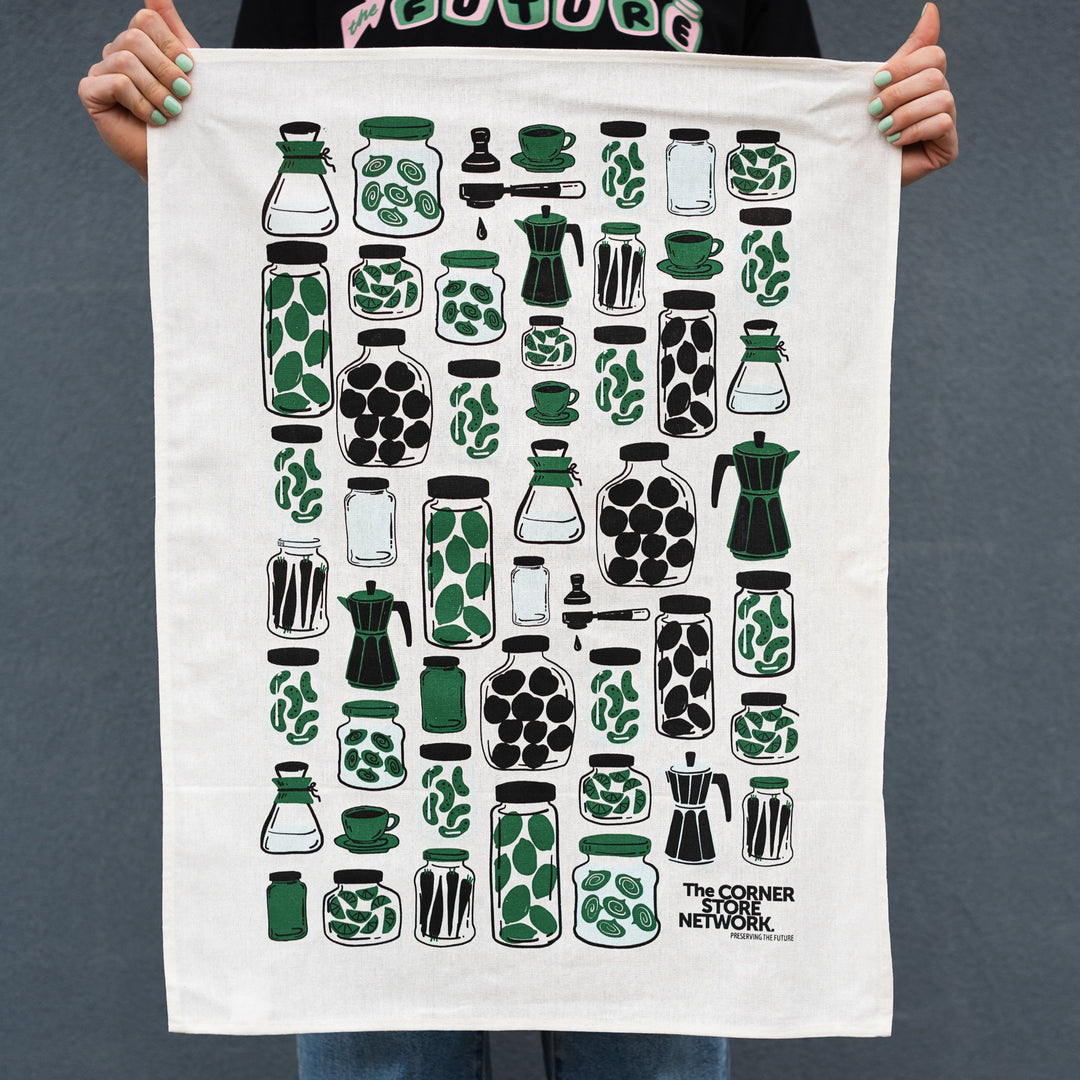 hands holding white linen and cotton blend tea towel with green, black and baby blue illustrated design showing various filled jars and coffee equipment