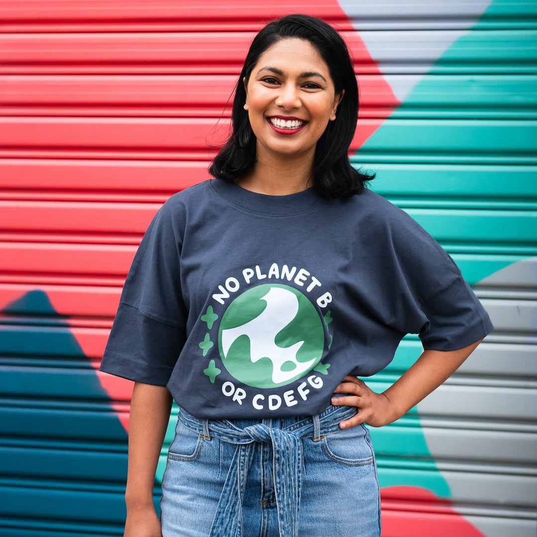 smiling woman in front of colourful wall wearing oversized grey organic cotton shirt with blue and green design saying no planet b or cdefg surrounding planet earth