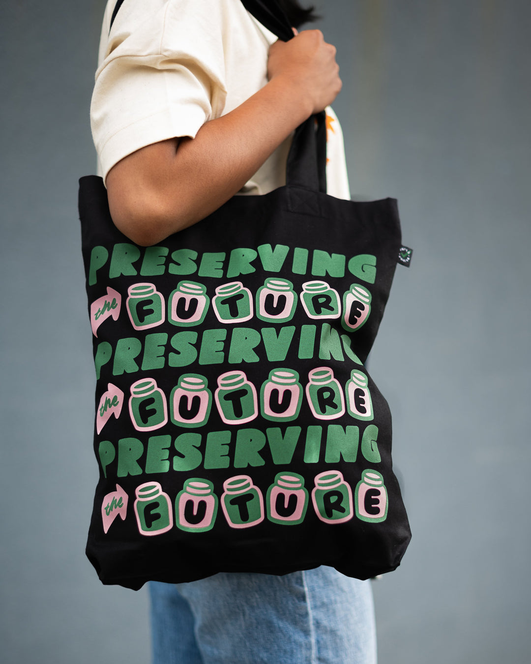 arm holding organic black cotton tote with pink and green design with three rows of green and pink text saying preserving the future