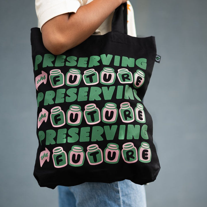 organic black cotton tote with pink and green design with three rows of text staying preserving the future, where the letters of future are in jars