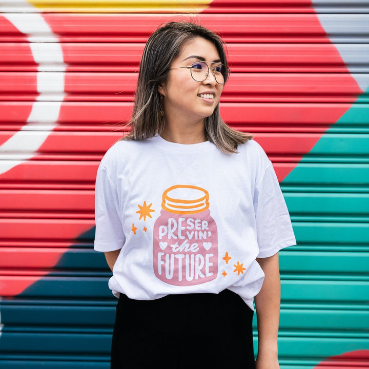 smiling woman in front of colourful wall wearing white organic cotton shirt with pink and orange design of a jar housing the words preserving the future