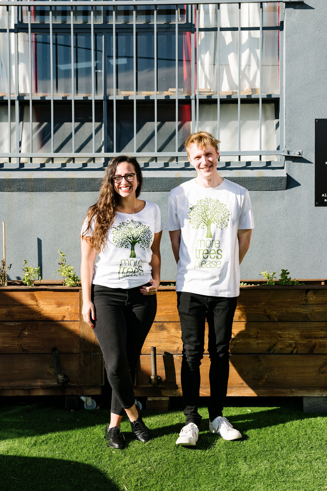 two smiling people wearing white organic cotton tees featuring a green illustrated design of a tree with the words more trees please underneath