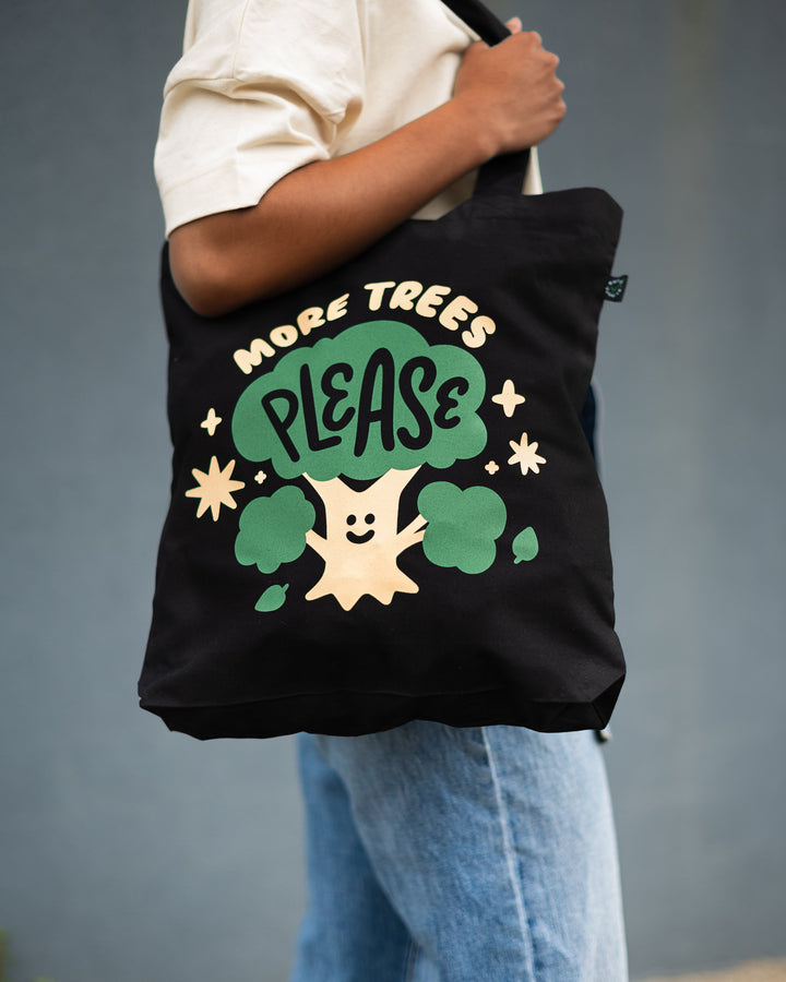arm holding organic black cotton tote with cream and green design showing a smiling tree with words more trees please