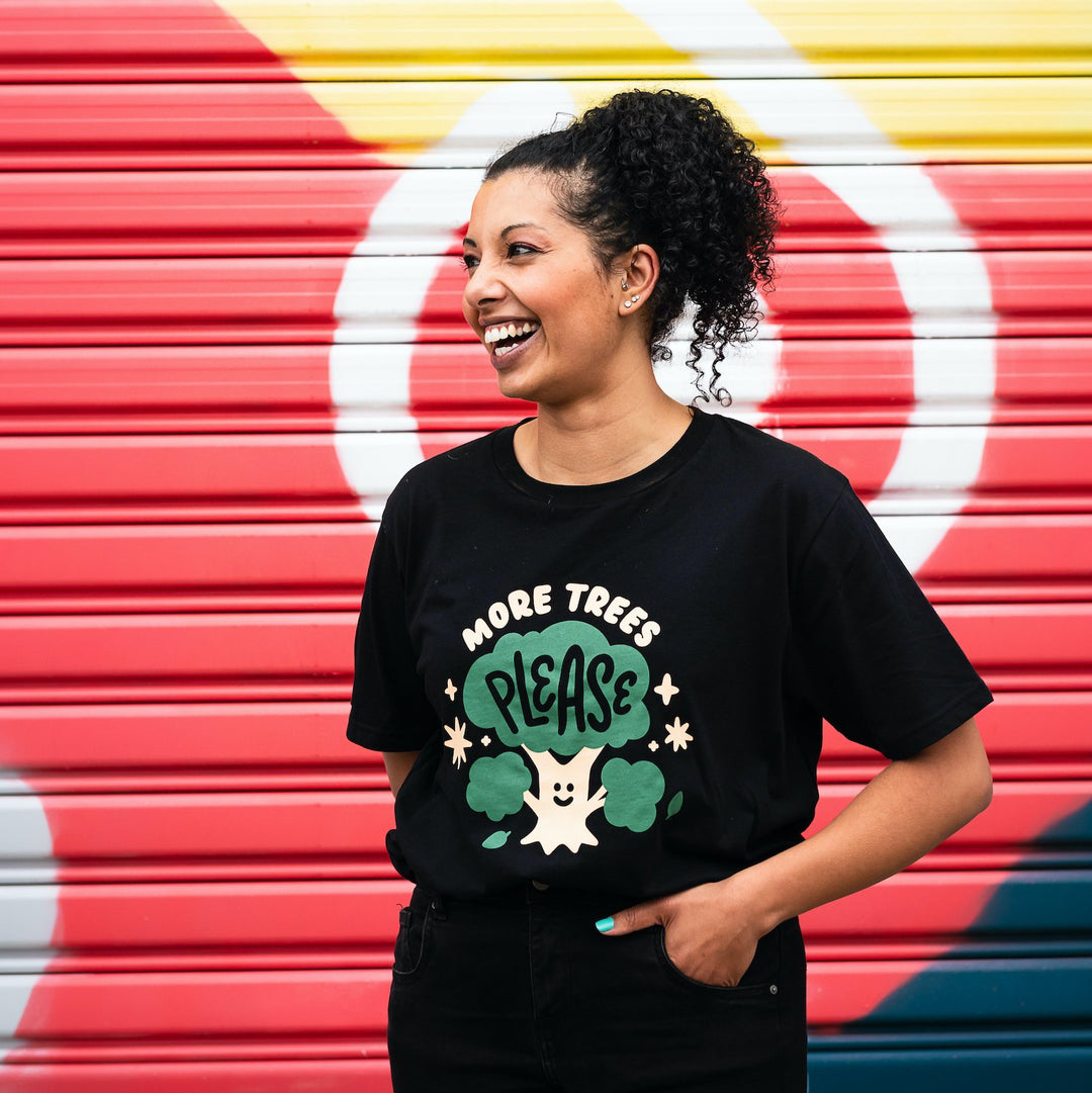 smiling woman in front of colourful wall wearing black organic cotton shirt with cute cream and green design showing a smiling tree with words more trees please