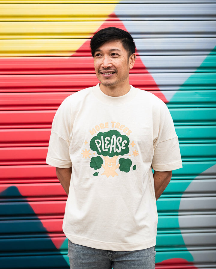 smiling man in front of colourful wall wearing oversized cream organic cotton shirt with cute cream and green design showing a smiling tree with words more trees please