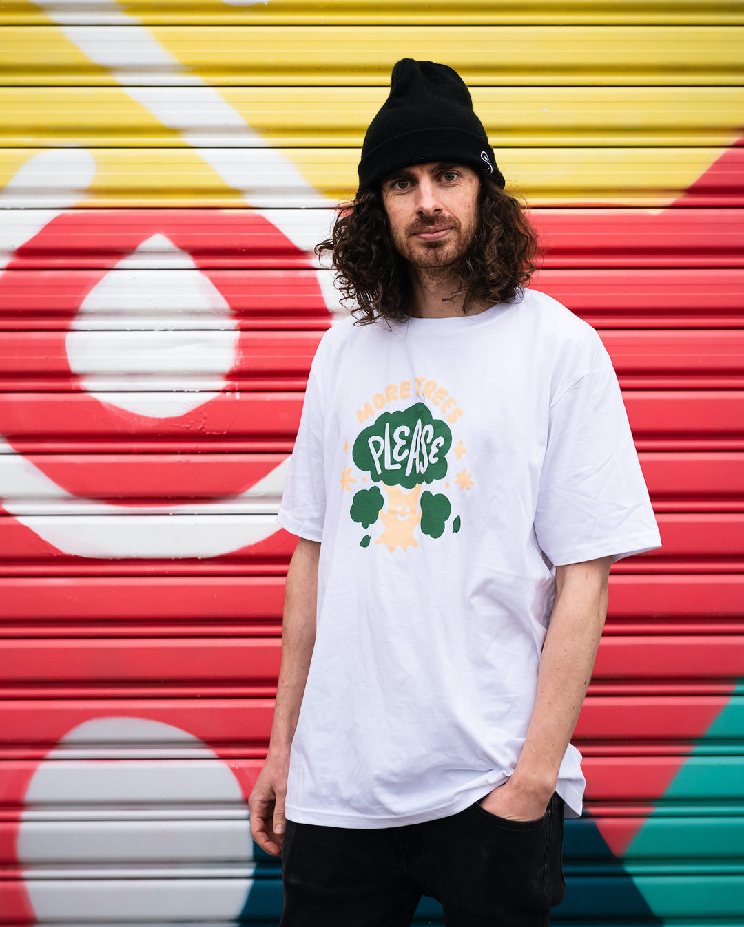 smiling man in front of colourful wall wearing white organic cotton shirt with cute cream and green design showing a smiling tree with words more trees please