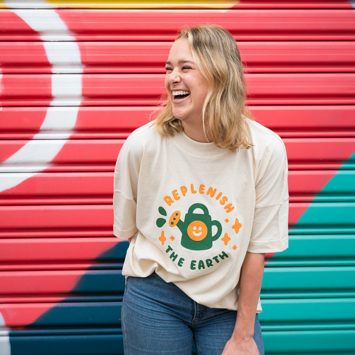 smiling woman in front of colourful wall wearing oversized cream organic cotton shirt with cute orange and green design of smiling watering can saying replenish the earth