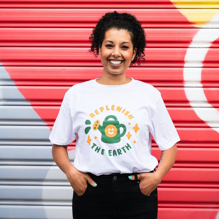smiling woman in front of colourful wall wearing white organic cotton shirt with cute orange and green design of smiling watering can saying replenish the earth